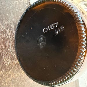 May include: Close-up of the bottom of a brown glass bottle. The base is dark brown with a circular design and the numbers "0157" and "914" printed in white. A circular logo is also visible.