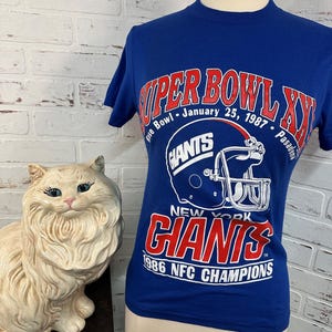 May include: Blue t-shirt with a red and white graphic of a football helmet and the text "Super Bowl XXI, January 25, 1987, Pasadena, Giants, New York Giants, 1986 NFC Champions".