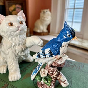 Op de afbeelding: Een keramieken beeldje van een blauwe gaai die op een tak zit met een kleine witte bloem. De vogel heeft blauwe, witte en zwarte markeringen. Op de achtergrond staat een wit kattenbeeldje. De beeldjes staan op een groene ondergrond.