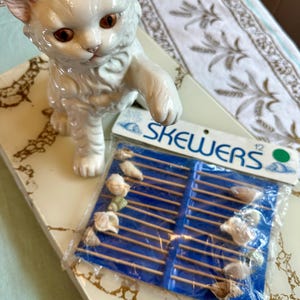 Puede incluir: Una figurita de gato de porcelana blanca con ojos marrones, de pie sobre una superficie de mármol. En primer plano, un paquete de 12 pinchos de madera con conchas marinas adheridas. El paquete es azul y blanco con la palabra "PINCHOS" impresa en él.