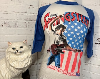 May include: Vintage white and blue baseball tee shirt with a graphic of Bruce Springsteen playing a guitar in front of an American flag. The text "Bruce Springsteen" and "Born in the USA '84" are printed on the shirt.
