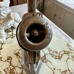 Puede incluir: Un antiguo Bates Automatic Eyeleter, una herramienta de metal con una cabeza circular y una palanca. La cabeza está marcada con el texto "AUTOMATIC EYELETER" y "THE BATES MANUFACTURING CO., ORANGE N.J.U.S.A." La herramienta es plateada y se encuentra sobre una superficie con un patrón dorado y blanco.