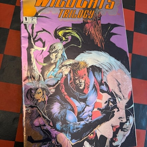 May include: A comic book titled "WildC.A.T.s Trilogy" with Issue #1. The cover features various illustrated characters in shades of purple, blue, red, and black. The comic book is set against a checkered background.