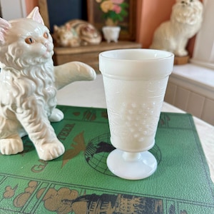 May include: A white milk glass pedestal glass with a textured pattern. The glass is on a green book with gold lettering and a world map design. Two white cat figurines are in the background.