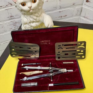 May include: A vintage Unitech Royal drafting set in a black case with a deep red velvet interior. The set includes a metal compass, two metal drafting templates with various cutouts, a small clear plastic container, and two black ink pens.