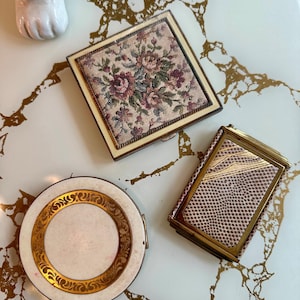 Op de afbeelding: Drie vintage compactpoeders op een wit en goud oppervlak. Een vierkante compactpoeder heeft een bloemen tapestry design. Een ronde compactpoeder heeft een gouden filigraan rand. De derde compactpoeder heeft een slangenpatroon.