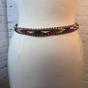 May include: A beaded belt with a red background and geometric patterns in white, black, yellow, and blue. The belt is displayed on a mannequin. The belt's design is a repeating pattern of triangles and rectangles.