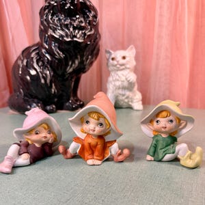 Könnte beinhalten: Drei Keramik-Elfenfiguren mit spitzen Hüten in Rosa, Orange und Gelb, die passende Outfits in Dunkellila, Orange und Grün tragen. Dahinter stehen eine große schwarze Katzenfigur und eine kleinere weiße Katzenfigur mit grünen Augen.