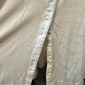 May include: Close-up of an off-white, button-down shirt. The shirt features a row of small, round buttons down the front and a detailed buttonhole seam. The fabric appears to be a soft, knit material.