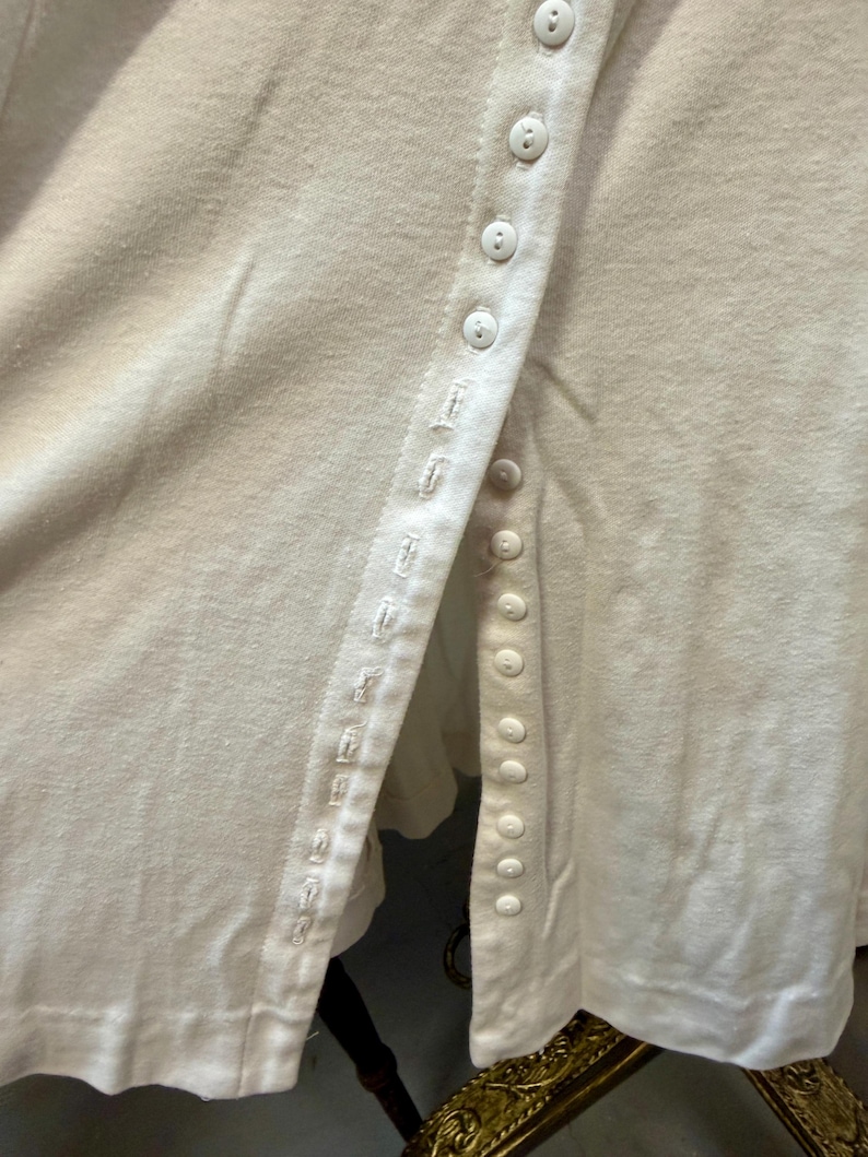 May include: Close-up of an off-white, button-down shirt. The shirt features a row of small, round buttons down the front and a detailed buttonhole seam. The fabric appears to be a soft, knit material.