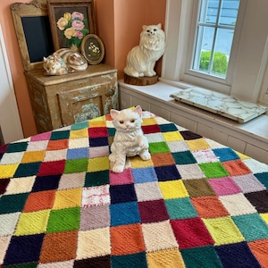 May include: A white ceramic cat figurine sits on a colorful patchwork quilt. The quilt features squares of various colors, including red, yellow, green, and blue. Other cat figurines and framed art are in the background.