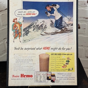 Könnte beinhalten: Gerahmte Vintage-Werbung für Borden's Hemo. Die Anzeige zeigt eine Skifahrerin, eine Zeichentrickkuh und Text, der für die Vitamine und Mineralien des Getränks wirbt. Die Anzeige enthält den Text "She's 35...Acts 25, That's My Hemo Gal!"