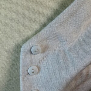 May include: Close-up of a light green knit sweater with a light pink cardigan. The cardigan has three white buttons. The sweater and cardigan are made of a soft, textured fabric.