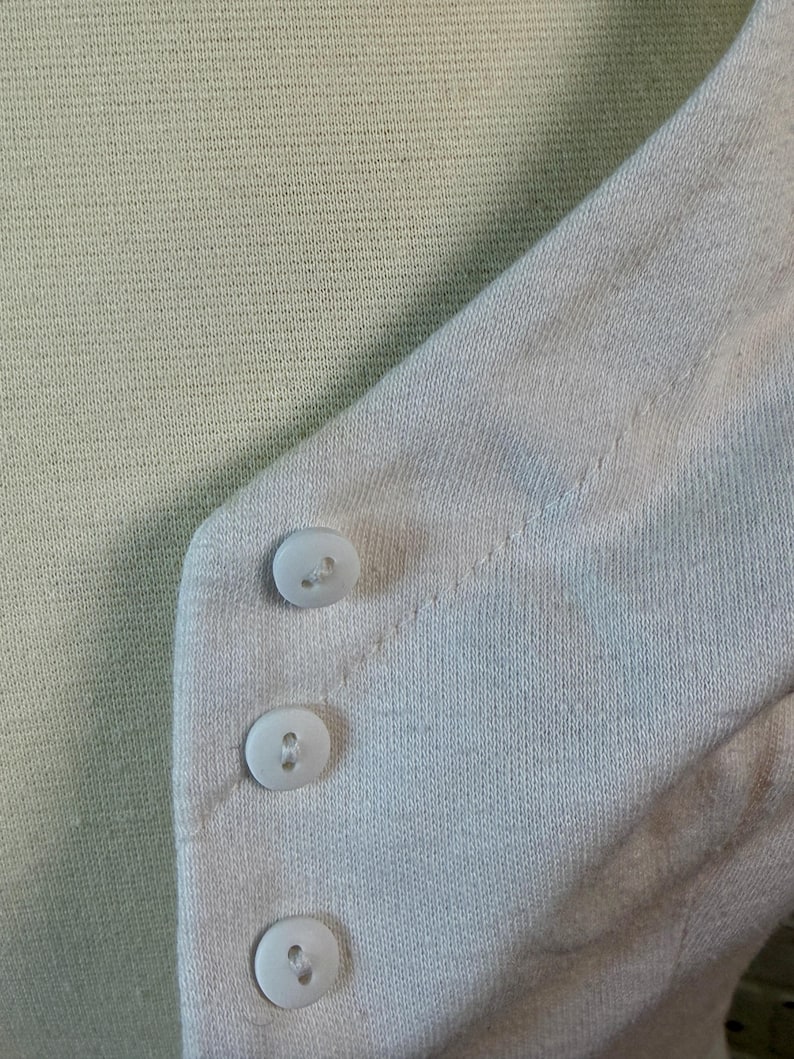 May include: Close-up of a light green knit sweater with a light pink cardigan. The cardigan has three white buttons. The sweater and cardigan are made of a soft, textured fabric.