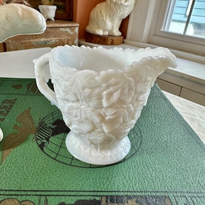 May include: A white, vintage creamer with a leaf design. The creamer has a handle and a spout, and sits on a round base. The creamer is made of a glossy, opaque material. The creamer is on a green surface.