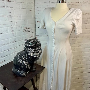 May include: A vintage white dress with short sleeves and a button-down front is displayed on a mannequin. A black ceramic cat statue with green eyes sits on a dark wooden side table. The dress has a V-neck and a fitted bodice.