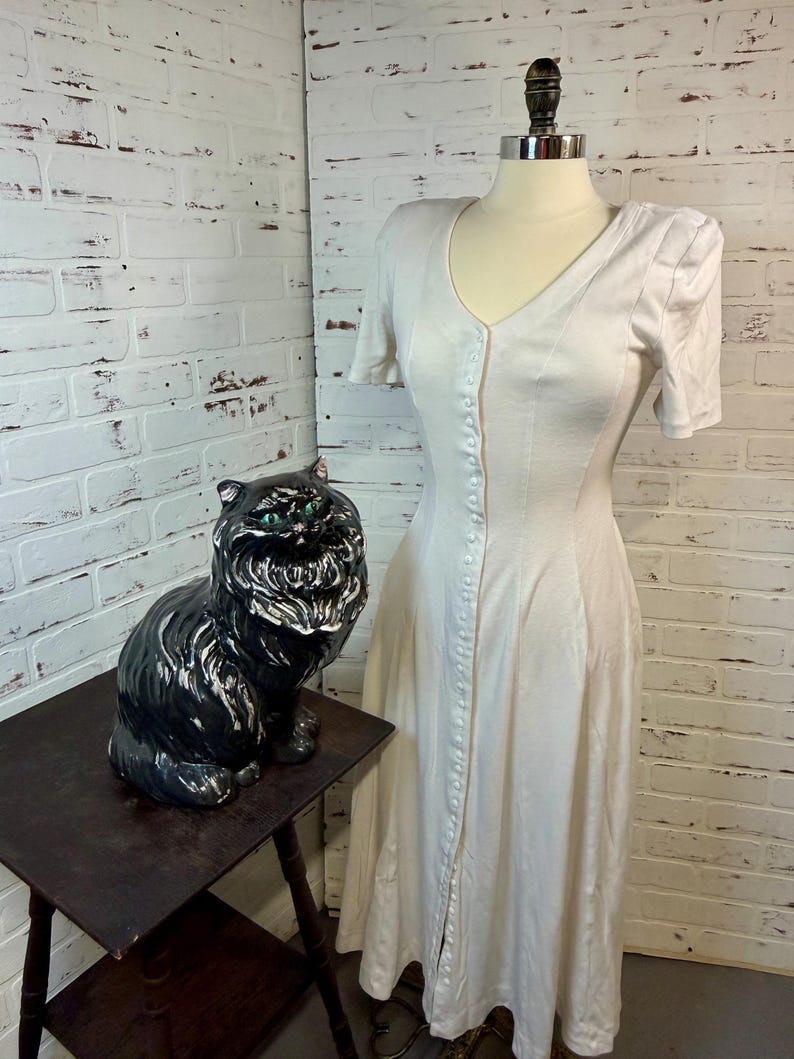 May include: A vintage white dress with short sleeves and a button-down front is displayed on a mannequin. A black ceramic cat statue with green eyes sits on a dark wooden side table. The dress has a V-neck and a fitted bodice.