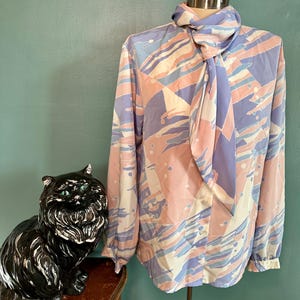 Puede incluir: Una blusa de manga larga con cuello de lazo. La blusa está hecha de una tela estampada de color rosa claro, azul y blanco. El lazo es de color morado claro.
