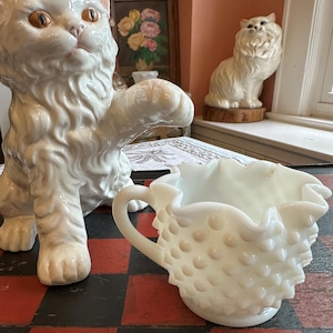 May include: A white ceramic cat figurine with orange eyes, sitting on a black and white checkered surface, next to a white milk glass pitcher with a ruffled rim and a textured surface.