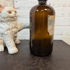 May include: A brown glass bottle with a black cap and a label that reads "SUPER". A white ceramic cat statue is to the left of the bottle. The bottle is on a dark brown surface.