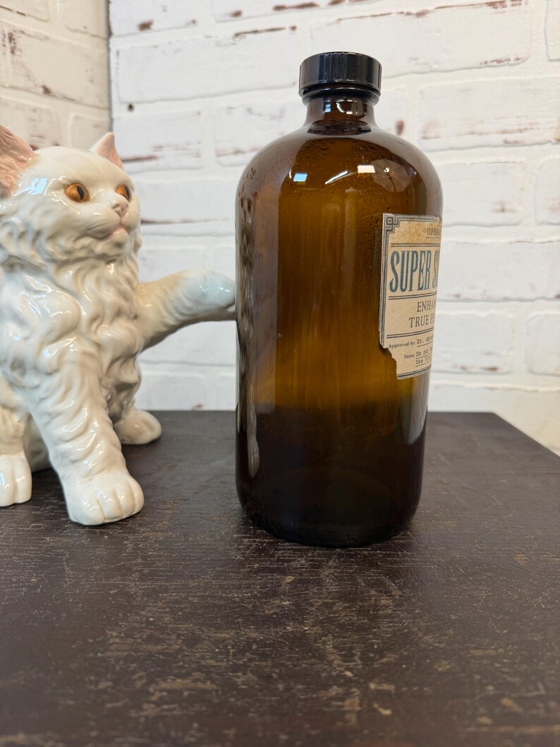 May include: A brown glass bottle with a black cap and a label that reads "SUPER". A white ceramic cat statue is to the left of the bottle. The bottle is on a dark brown surface.