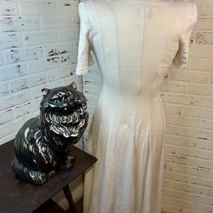 May include: A cream-colored dress with short sleeves and a fitted bodice is displayed on a mannequin. A black ceramic cat statue with green eyes sits on a dark wooden table. The background features a white brick wall.