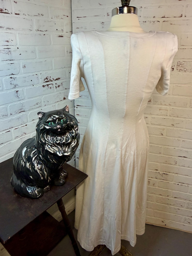 May include: A cream-colored dress with short sleeves and a fitted bodice is displayed on a mannequin. A black ceramic cat statue with green eyes sits on a dark wooden table. The background features a white brick wall.