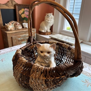 May include: A white porcelain cat figurine sitting inside a brown and beige woven wicker basket with a wooden handle.