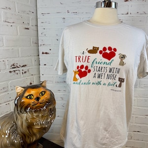 May include: White t-shirt with the text "A true friend starts with a wet nose and ends with a tail." The shirt features paw prints, hearts, and cartoon animal graphics. A ceramic cat statue is in the background.