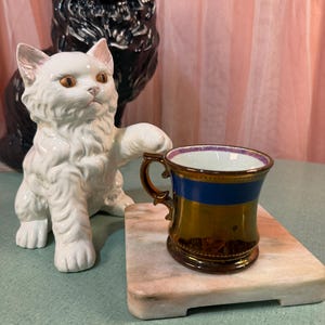May include: A white ceramic cat figurine with orange eyes and a raised paw positioned next to a small, metallic gold and blue lusterware cup. The cup features a dark gold base, a horizontal blue band, and a decorative rim with a purple pattern.