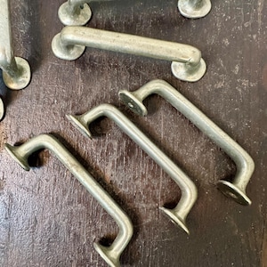 May include: A collection of antique metal drawer pulls. The pulls are a muted silver color, with a curved handle and circular mounting plates. The hardware is likely made of cast iron or a similar metal. The pulls are designed for cabinets or drawers.