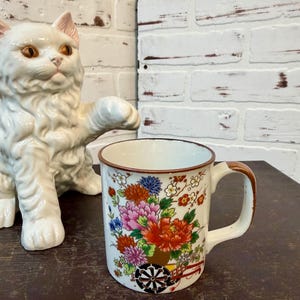 May include: A ceramic mug with a brown rim and handle, decorated with a vibrant floral design in shades of red, pink, blue, and yellow. The mug features a decorative cartwheel design at the base. A white ceramic cat statue is in the background.