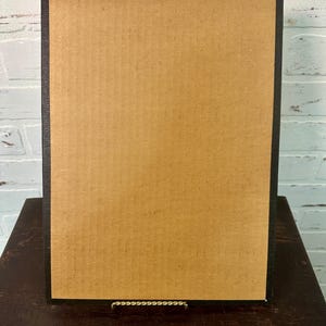 Może przedstawiać: Prostokątne czarne oprawione dzieło sztuki z brązowym kartonowym tyłem. Grafika jest wyświetlana na małym metalowym sztaludze, na tle białej ceglanej ściany i ciemnej drewnianej powierzchni.