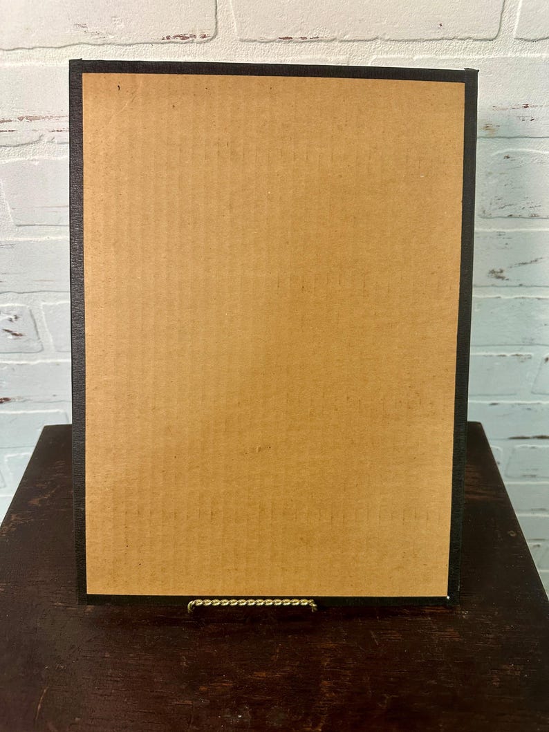 Może przedstawiać: Prostokątne czarne oprawione dzieło sztuki z brązowym kartonowym tyłem. Grafika jest wyświetlana na małym metalowym sztaludze, na tle białej ceglanej ściany i ciemnej drewnianej powierzchni.