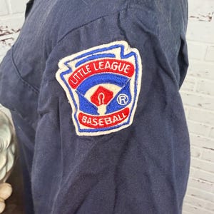 May include: A dark navy blue long-sleeved shirt featuring an embroidered Little League Baseball patch on the upper sleeve. The patch is shaped like a home plate with red, white, and blue stitching, displaying the text Little League Baseball.