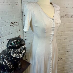 May include: A light-colored, button-down dress with short sleeves is displayed on a mannequin. The dress has a V-neck and a row of buttons down the front. A black ceramic cat statue with green eyes sits to the left of the dress.