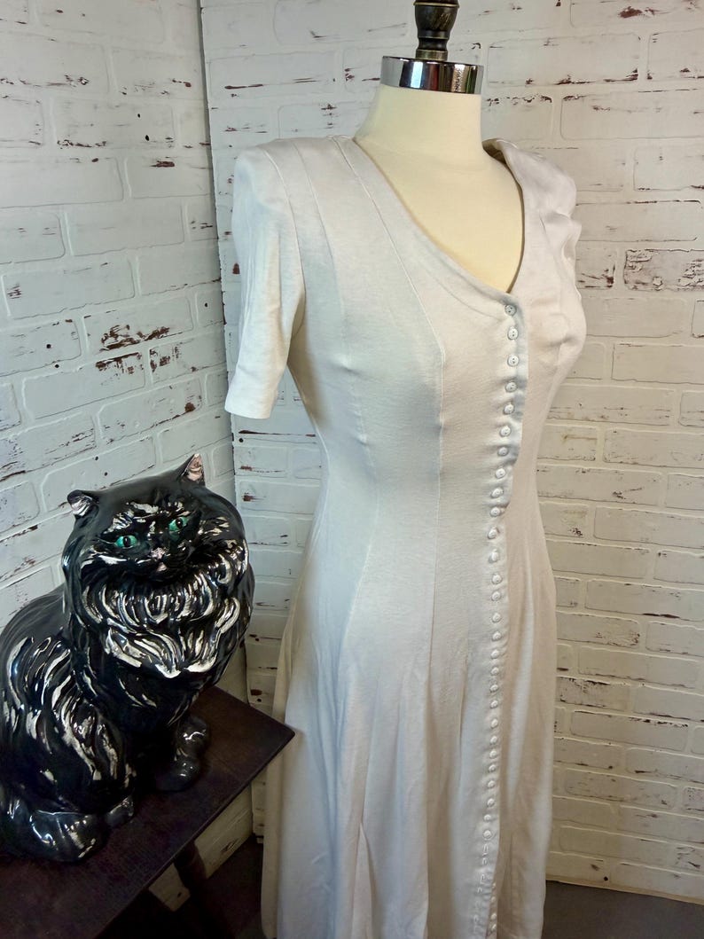 May include: A light-colored, button-down dress with short sleeves is displayed on a mannequin. The dress has a V-neck and a row of buttons down the front. A black ceramic cat statue with green eyes sits to the left of the dress.