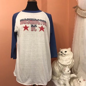 Puede incluir: Una camiseta de béisbol vintage blanca y azul con el texto "WASHINGTON DC" en rojo, blanco y azul con dos estrellas rojas. La camiseta tiene un diseño de bloques de color azul y blanco.
