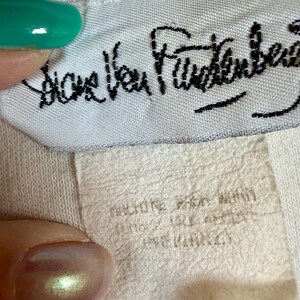 May include: Close-up of a white fabric tag with the black cursive text "Diane Von Furstenberg". Below the tag is a smaller, off-white tag with faded text. The image is of a clothing label.