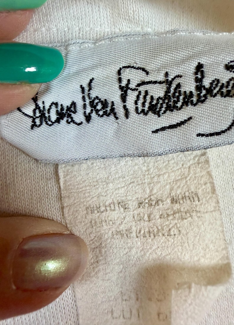 May include: Close-up of a white fabric tag with the black cursive text "Diane Von Furstenberg". Below the tag is a smaller, off-white tag with faded text. The image is of a clothing label.