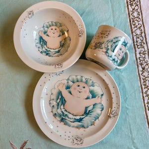 May include: A white ceramic children's dish set featuring a baby design. The set includes a bowl, a plate, and a mug. The baby is surrounded by a blue and green design. The plate and bowl have a white border with small bunny illustrations.