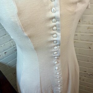 May include: A light-colored, button-down dress with a fitted silhouette. The dress features a series of small, white buttons running down the front and a subtle seam detail. The fabric appears to be a soft, knit material.