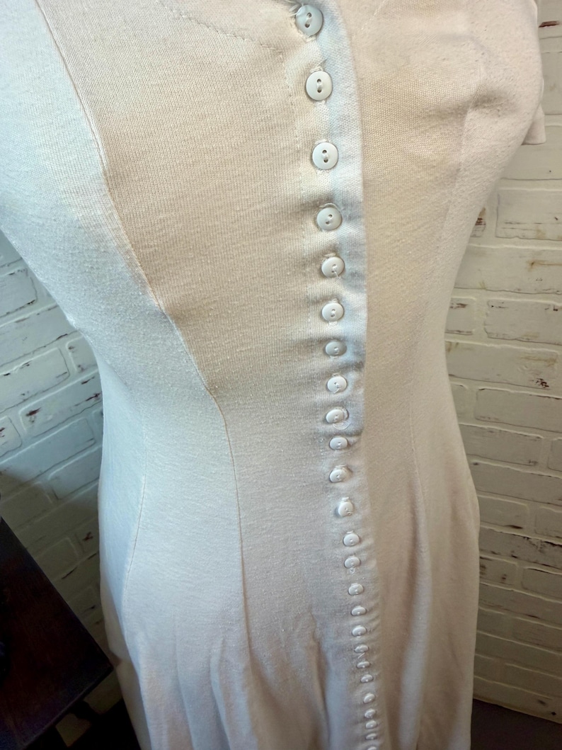 May include: A light-colored, button-down dress with a fitted silhouette. The dress features a series of small, white buttons running down the front and a subtle seam detail. The fabric appears to be a soft, knit material.