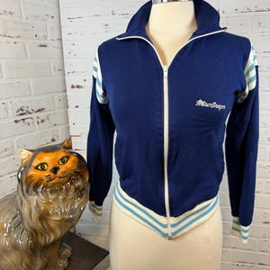 May include: Navy blue zip-up track jacket with white and light blue stripes on the sleeves and hem. The jacket has a stand-up collar and the text "MacGregor" embroidered on the chest. The jacket is displayed on a mannequin.