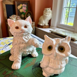 May include: A white ceramic cat figurine with orange eyes and a raised paw sits next to a white owl figurine with large yellow eyes. Both are on a green book. Other cat figurines are in the background.