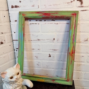 Puede incluir: Un marco de fotos rectangular con un acabado colorido y desgastado. El marco está pintado en tonos de verde, rojo, azul y amarillo. El marco está vacío y listo para una foto o una obra de arte. Una figura de gato de cerámica blanca está en primer plano.