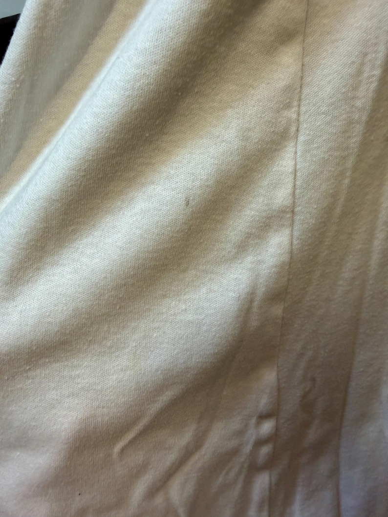 May include: Close-up of a white fabric with a textured surface. The fabric is slightly wrinkled, with a vertical seam running down the center. The material appears to be a soft, possibly knit, textile.