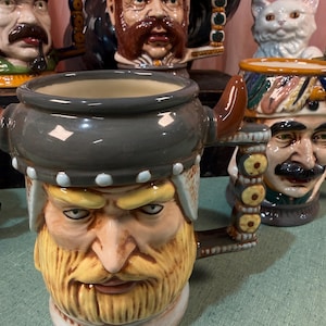 May include: A ceramic character mug shaped like a bearded person wearing a grey helmet with a horn. The handle features a vertical row of yellow circular accents. The mug has a detailed facial expression with blonde hair and a matching beard.