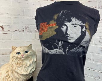 May include: Black sleeveless t-shirt with a faded image of Bruce Springsteen on the front. The text "Bruce Springsteen Born in the U.S.A." is printed on the shirt.