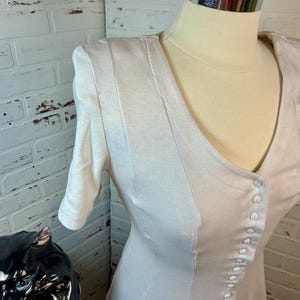 May include: A cream-colored dress with a V-neckline and short sleeves. The dress features a row of small buttons down the front and vertical seams for a tailored look. The dress is displayed on a mannequin.